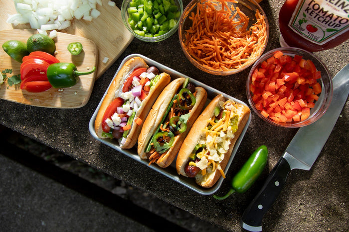 Three gourmet got dogs on a picnic table surrounded by ingredients