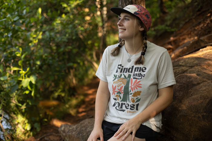 Woman sitting on a rock wearing a Find me in the Forest tee