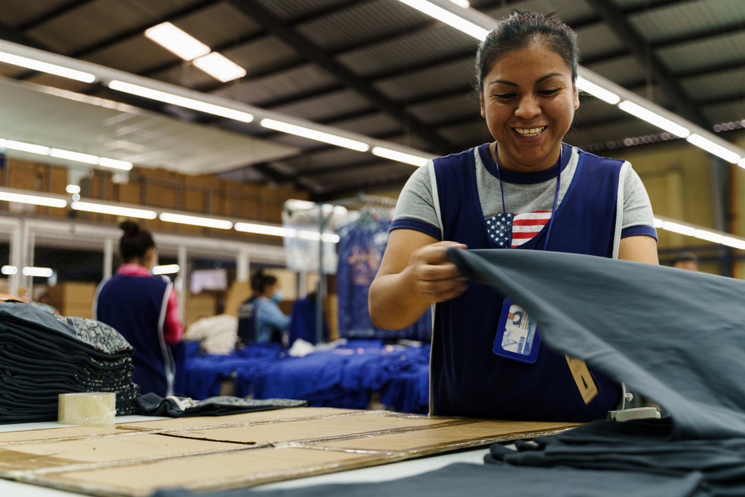 Woman smiling as she makes a Landmark Project tee shirt in a factory. Other workers in the background. 